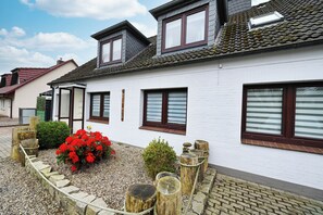 Une maison blanche avec un toit en pente, de nombreuses fenêtres et un jardin agrémenté de fleurs rouges et d’une clôture en bois.
