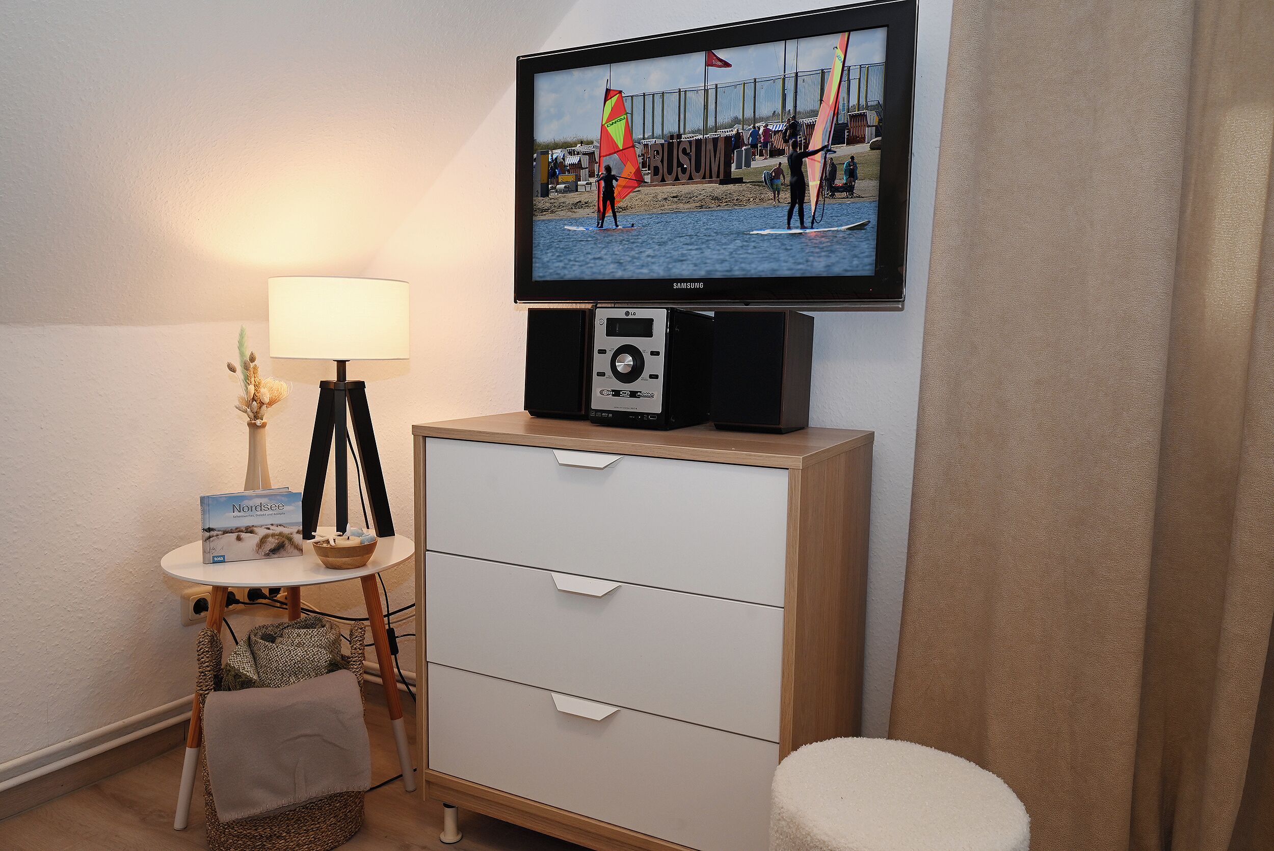 A hotel room with a television mounted on the wall, a bedside table with a lamp, a small chair, and a wooden dresser.