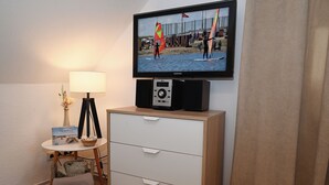 A hotel room with a television mounted on the wall, a bedside table with a lamp, a small chair, and a wooden dresser. - Noah's Ark - House by the sea<br>Apartment Koog (Büsum)