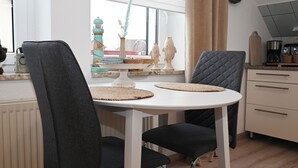 A dining area with a white table, two black chairs, and a window with decorative items. - Noah's Ark - House by the sea<br>Apartment Koog (Büsum)