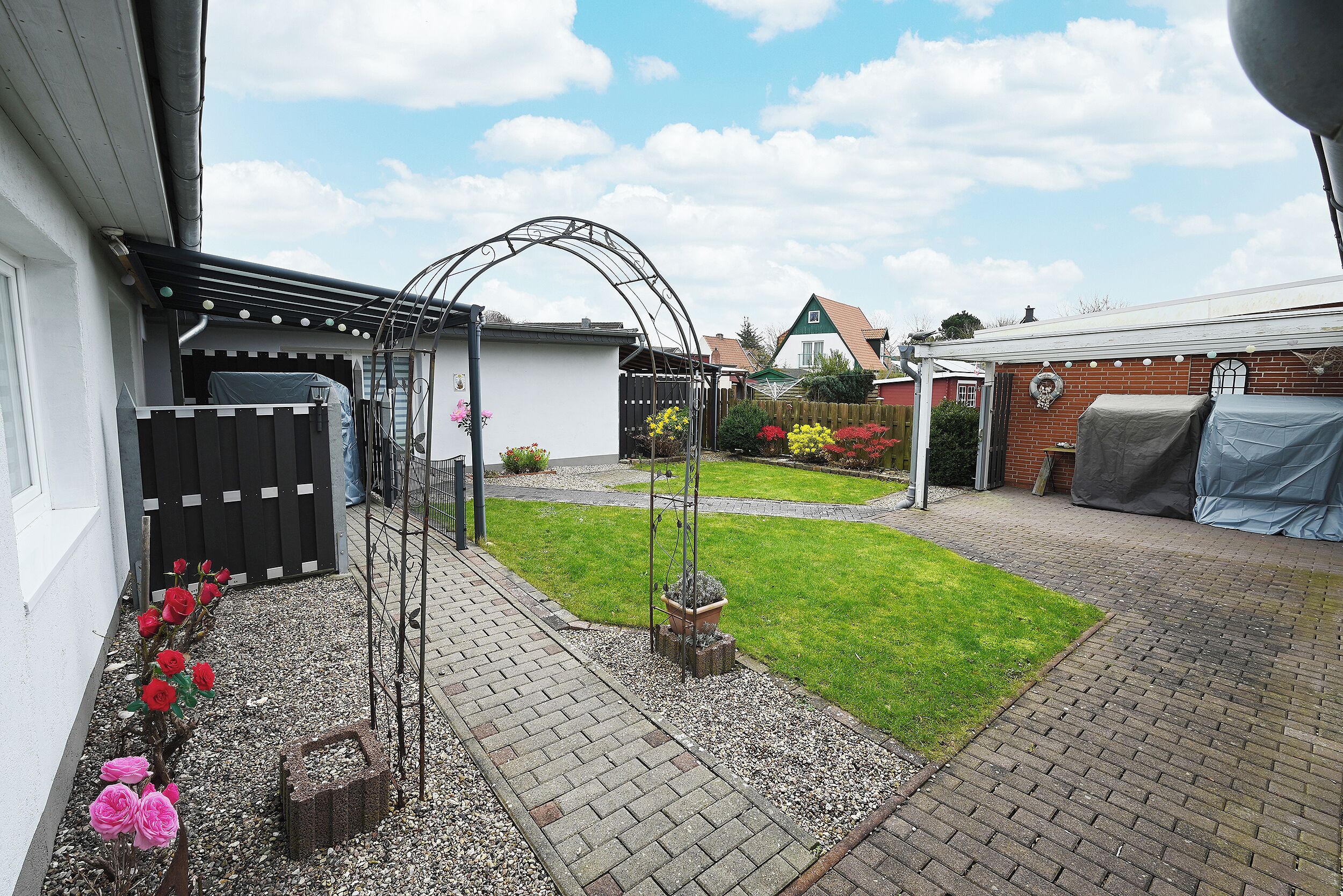 A backyard with a paved patio, a small garden with flowers, and a metal archway.