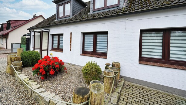 A white house with a sloped roof, multiple windows, and a garden with red flowers and a wooden fence. - Noah's Ark - House by the sea<br>Apartment Sea (Büsum)