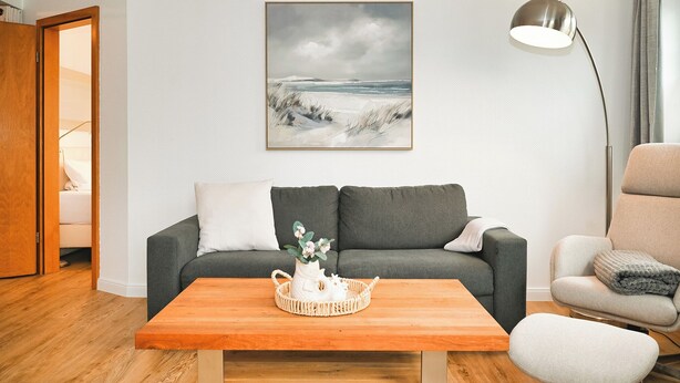 Una sala de estar con un sofá gris, una mesa de centro de madera, una lámpara de pie y un cuadro con una playa.