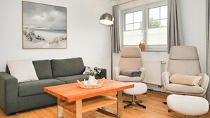 Un salón con un sofá gris, una mesa de centro de madera y dos sillones. Un cuadro de una playa adorna la pared, y se ve una ventana con cortinas.