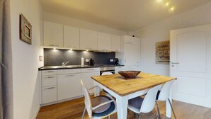 Cocina compacta con muebles blancos, una mesa de comedor de madera y una puerta blanca.