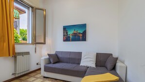 Una habitación con un sofá gris, una manta amarilla, un radiador, una ventana con cortinas y un cuadro de un paisaje urbano en la pared.