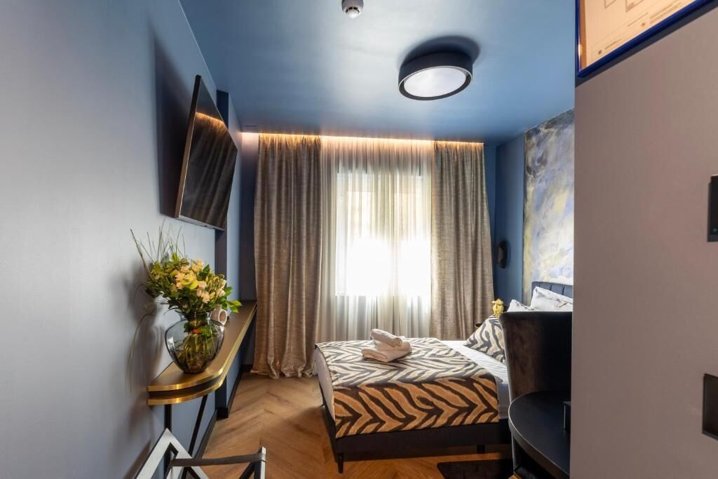 A hotel room with a bed, a desk, a chair, a TV, a window with curtains, and a wall-mounted artwork.