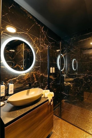 Ein modernes Badezimmer mit Marmor-Spritzschutz, einem Holz-Waschtisch, einem großen, ovalen Waschbecken und zwei wandmontierten Spiegeln mit runden Leuchten.