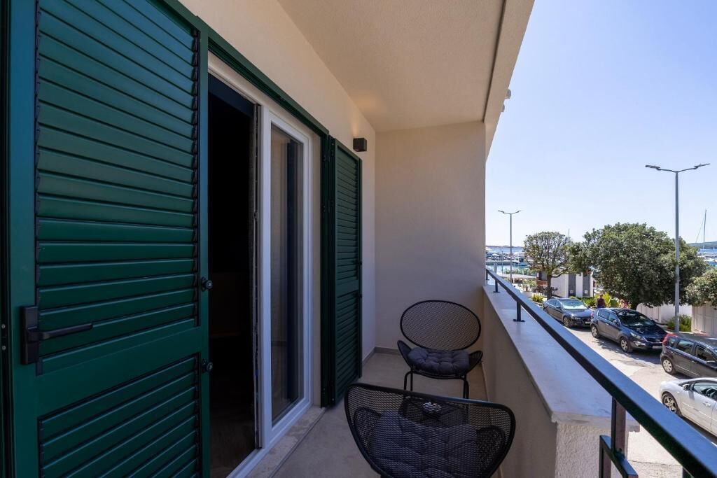 Un balcon avec une chaise, une porte avec des volets verts, et une vue sur les voitures garées et les arbres.