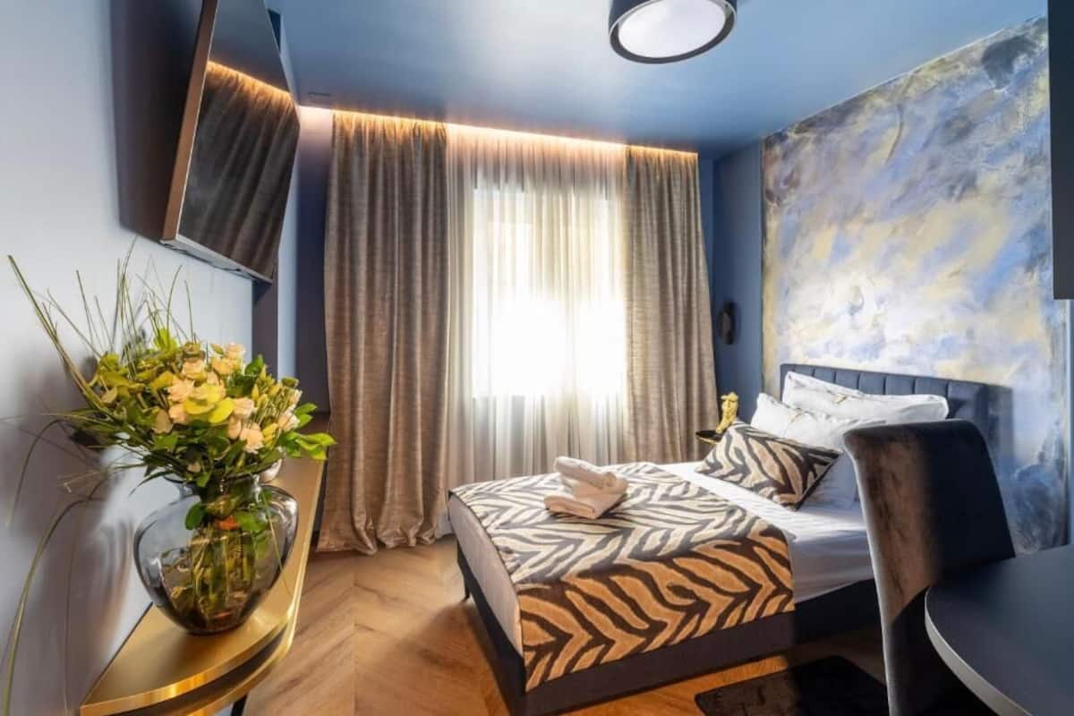 Ein modernes Hotelzimmer mit Bett, Schreibtisch, Stuhl, Blumen in einer Vase und einem großen abstrakten Gemälde an der Wand.