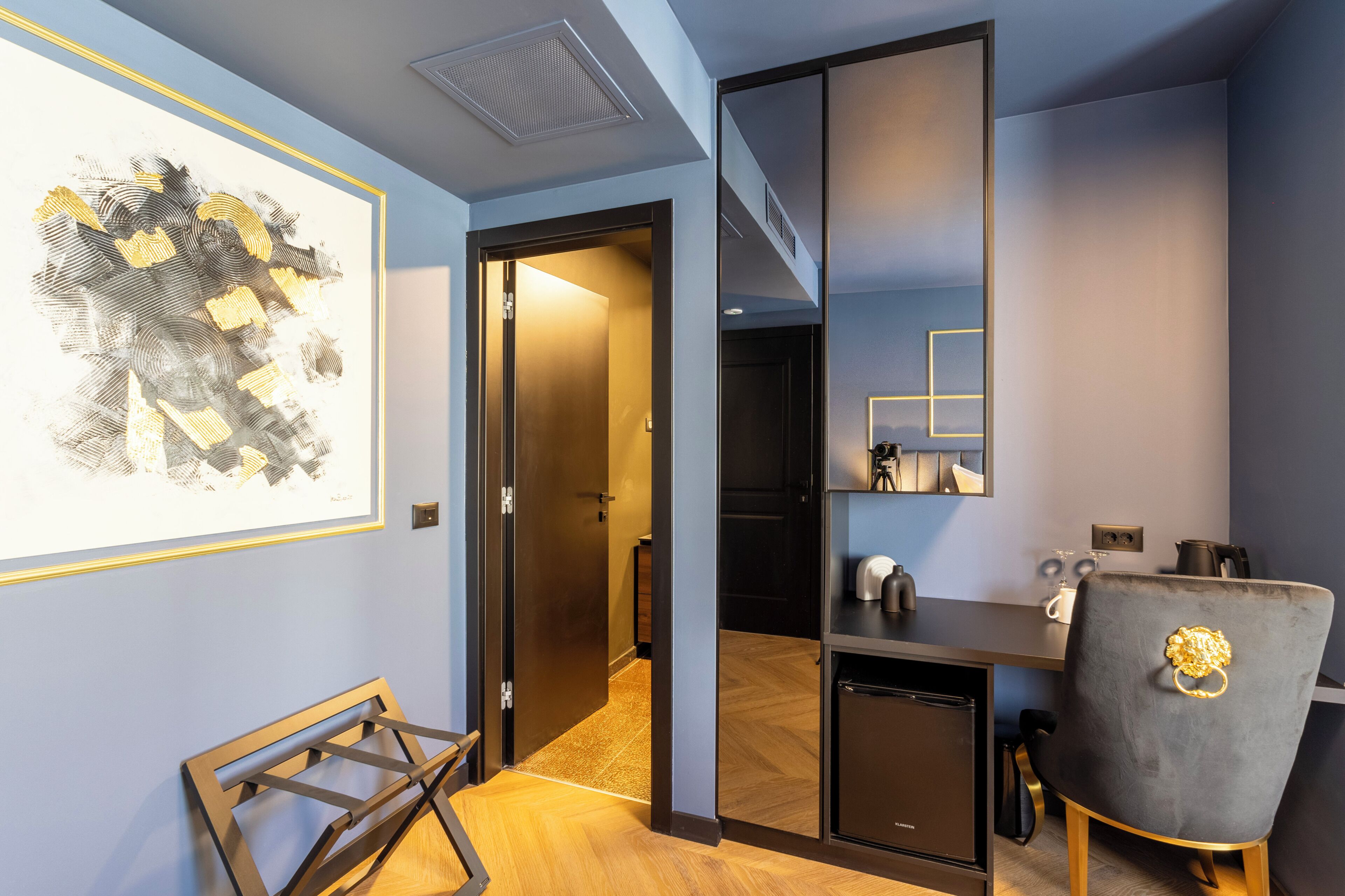 A modern hotel room with a wooden floor, a desk with a chair, a small refrigerator, and a framed artwork on the wall.