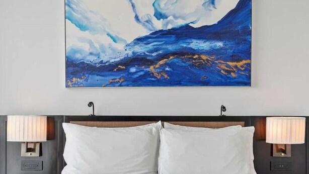 Een hotelkamer met een groot abstract schilderij aan de muur, een bed met witte lakens, en twee bedlampjes.