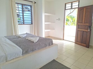 Een kamer met een bed, een raam met zonwering, een deur en uitzicht op groen.