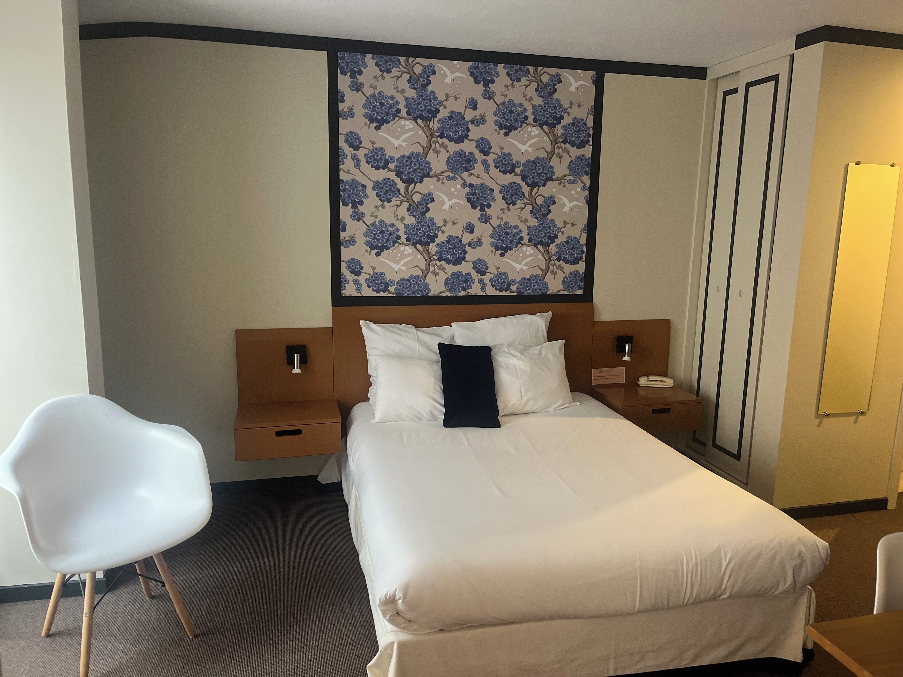 Ett hotellrum med en stor säng, en vit stol, ett sängbord, ett litet bord vid sängen och ett konstverk som hänger på väggen.