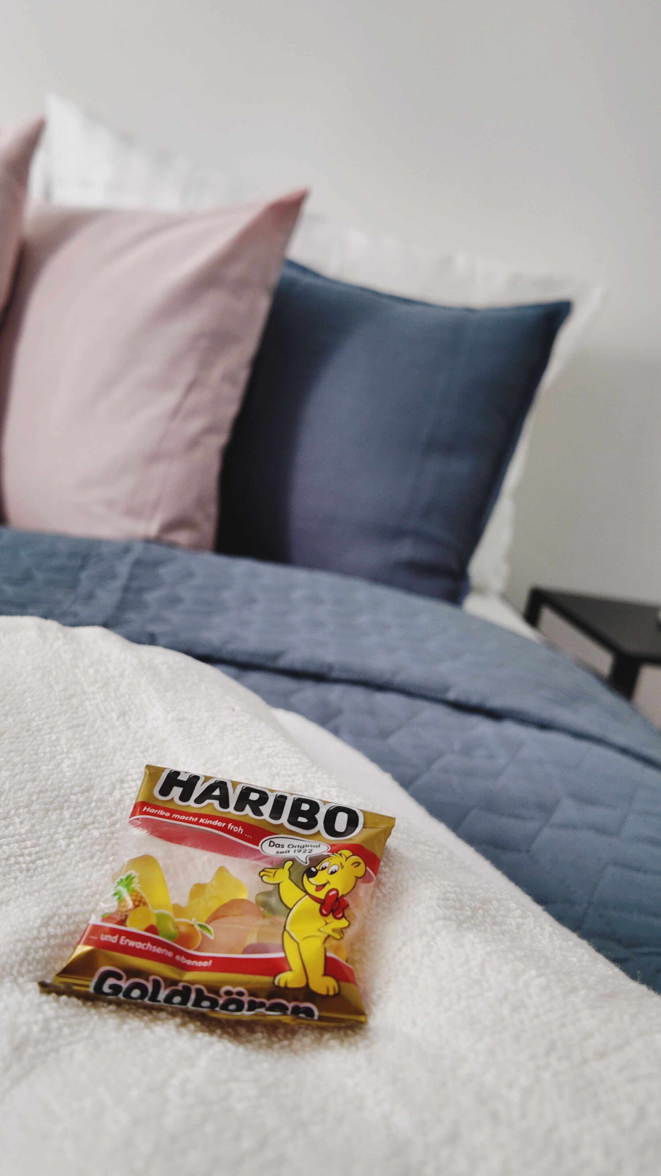 Ein Bett mit blauer Decke und Kissen, ein Paket Haribo Goldbären auf dem Bett.