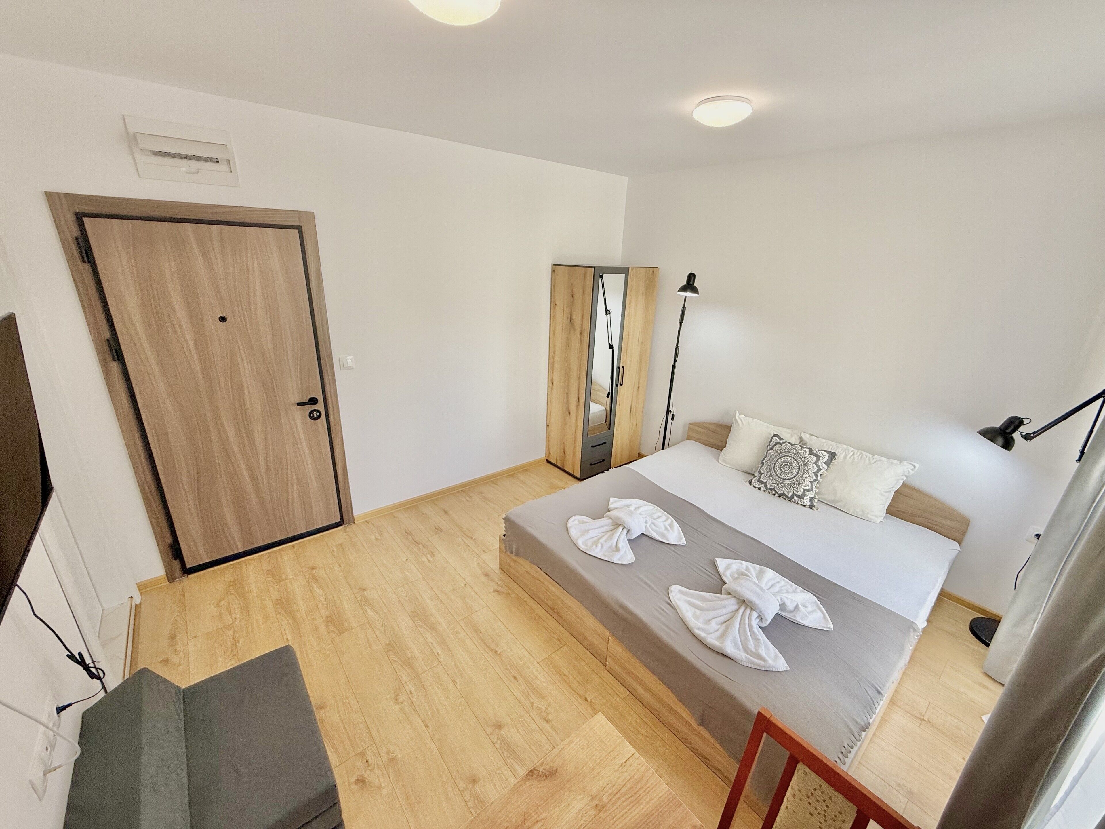A hotel room with a wooden floor, a bed with white linens and decorative pillows, a wardrobe, a desk with a chair, and a ceiling light.
