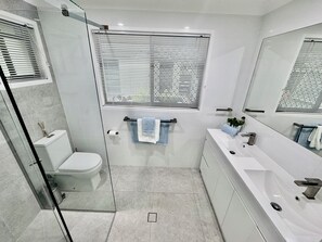 Ein modernes Badezimmer mit Duschkabine aus Glas, Toilette, Waschbecken mit Spiegel und Fenster mit Jalousien.