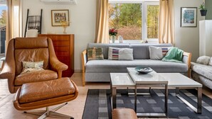 Een gezellige woonkamer met een leren fauteuil, een grijze bank, een salontafel en een raam met gordijnen.