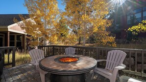 An outdoor patio with a round table and two Adirondack chairs, surrounded by a wooden deck and a stone fire pit.