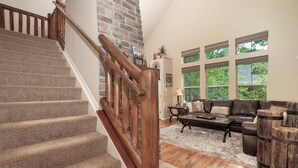 A staircase with wooden railings leading to a living room with a leather sofa, wooden coffee table, and large windows offering a view of greenery outside.