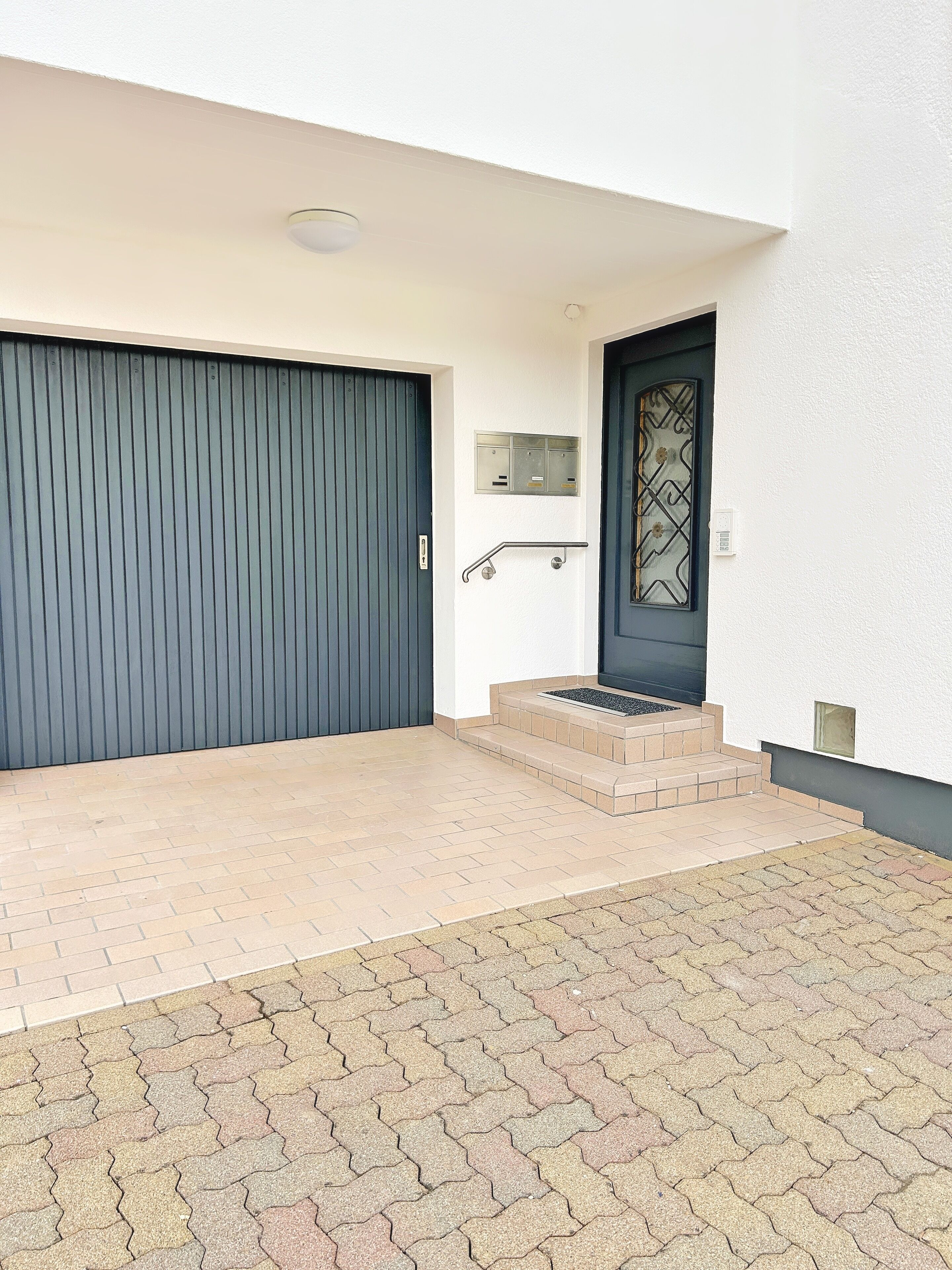 Une entrée moderne avec un portail de garage gris fermé, une porte noire agrémentée d’un panneau de verre décoratif et un petit escalier beige menant à l’entrée.