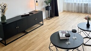 Una sala moderna con un televisor de pantalla plana, suelo de madera oscura, una consola negra, dos mesas redondas con copas de vino y cortinas blancas traslúcidas.