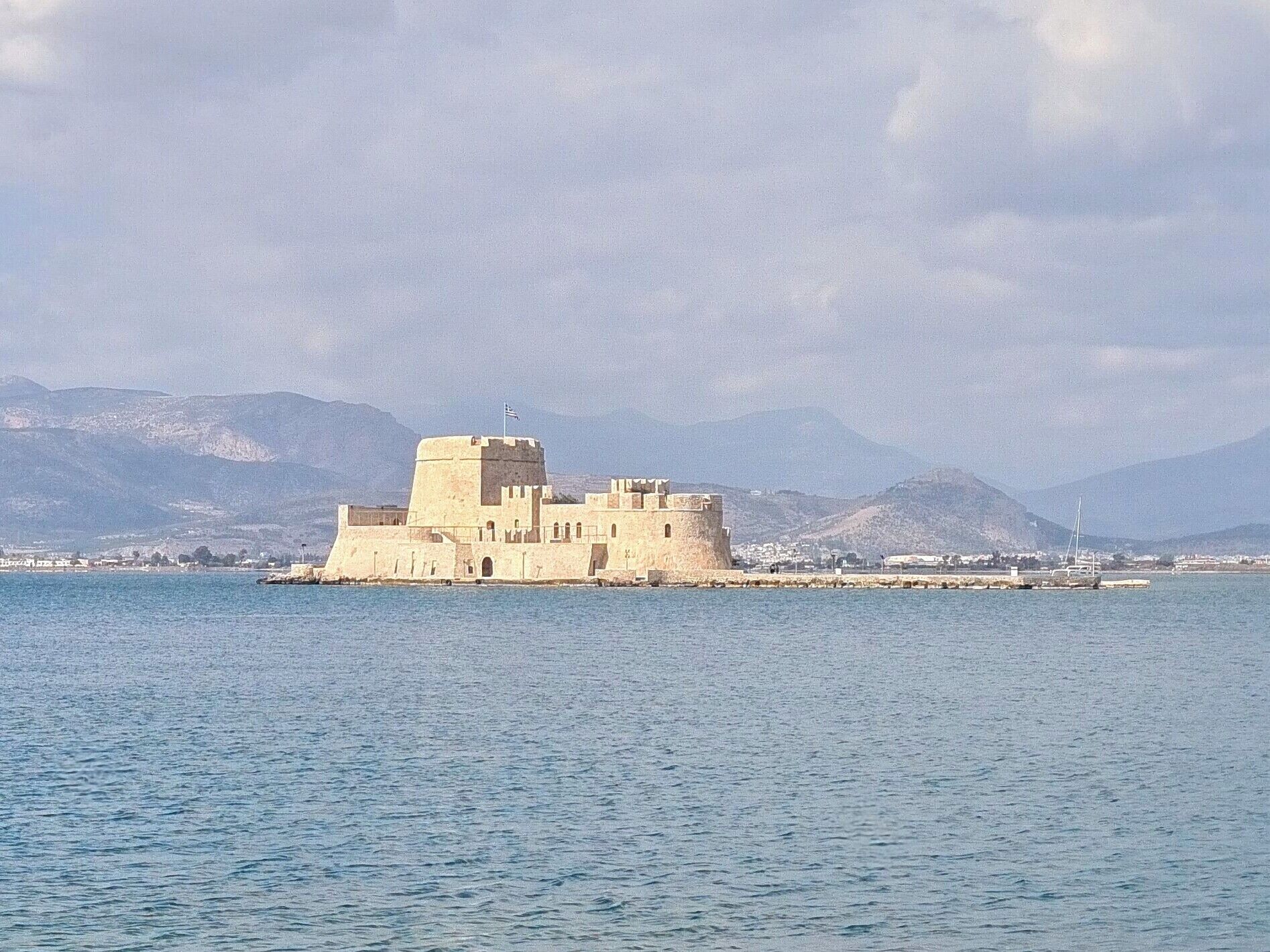A fortified building with multiple towers, situated on a small island in a body of water, with mountains in the background.