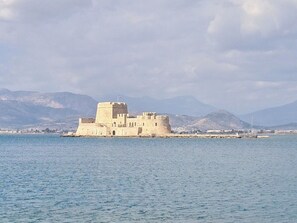 A fortified building with multiple towers, situated on a small island in a body of water, with mountains in the background.