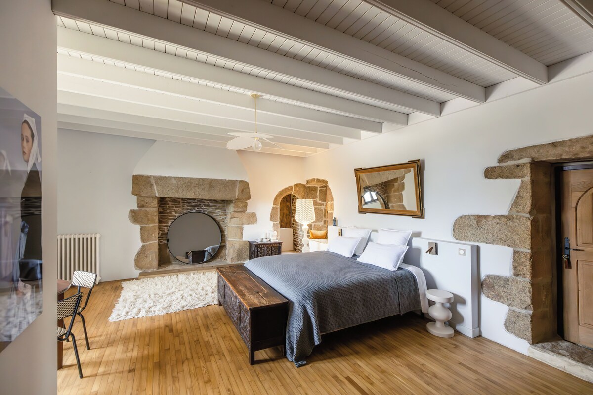 Un dormitorio con chimenea de piedra, techo de madera, cama con ropa blanca de cama y suelo de madera.
