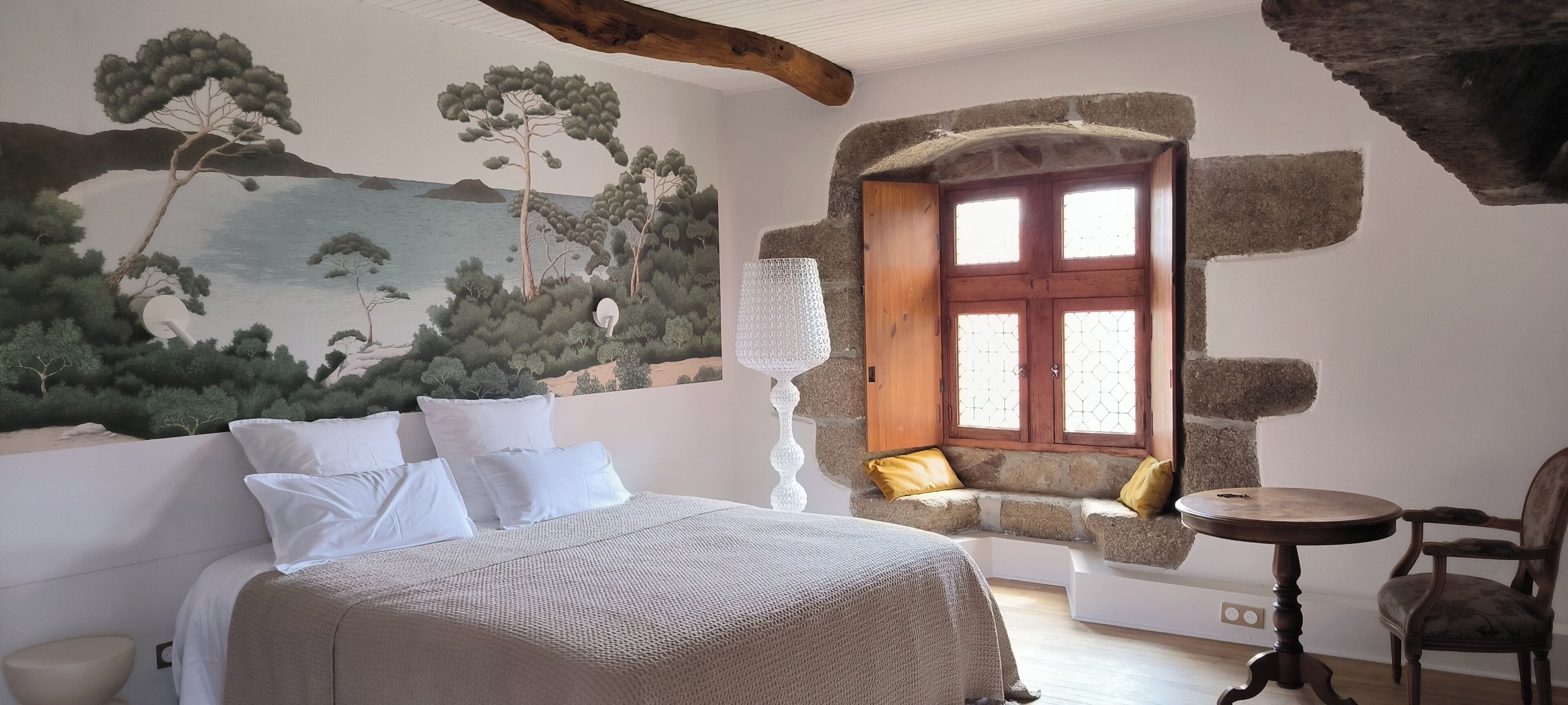 A bedroom with a bed, a stone bench with cushions, a table, and a window with a view of the sea.