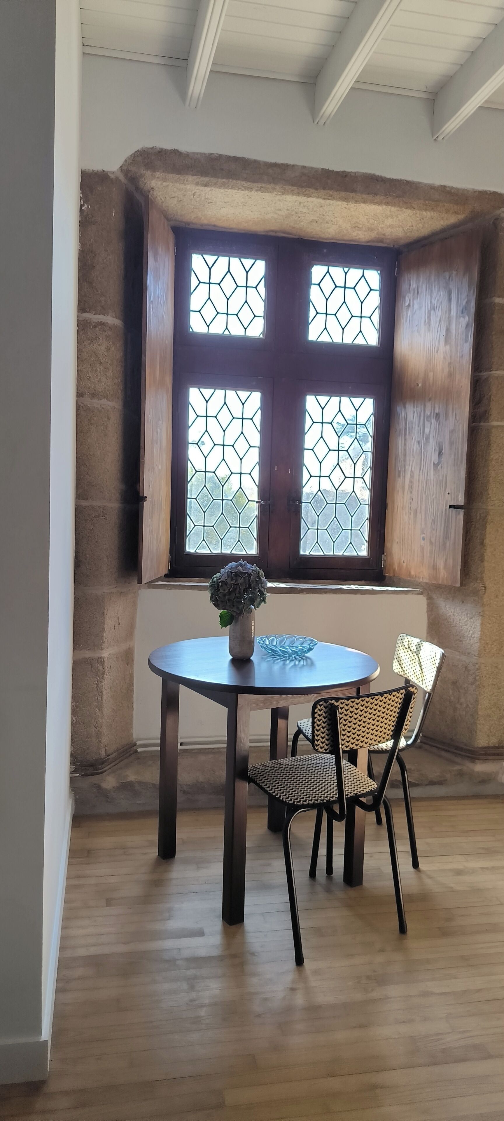 Una habitación de esquina con una mesa y sillas de madera, una ventana con diseño geométrico y una pared de piedra.