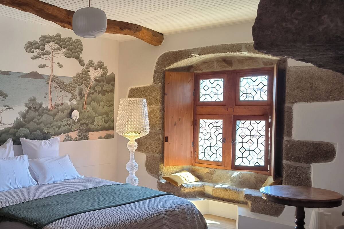 Ein Schlafzimmer mit einem Bett, einer Steinwand, einem Fenster mit einer Holztür und einem Wandgemälde.