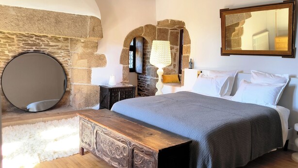 A bedroom with a stone wall, a round mirror, a bed with white linens, a wooden bench, and a small table with a lamp. - Manoir de Kerenneur (Plourin)