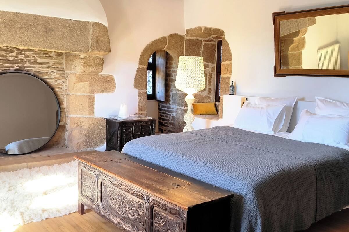 Ein Schlafzimmer mit Stehwänden, einem runden Spiegel, einem Bett mit weißer Bettwäsche, einer Holzbank und einem kleinen Tisch mit Lampe.