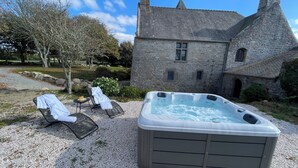 A hot tub and lounge chairs in a garden area with a stone building in the background. - Manoir de Kerenneur (Plourin)