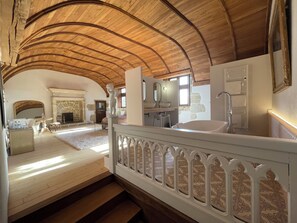 Una habitación amplia con un techo alto de madera, una chimenea de piedra, un ventanal grande y un baño moderno con bañera y lavamanos.