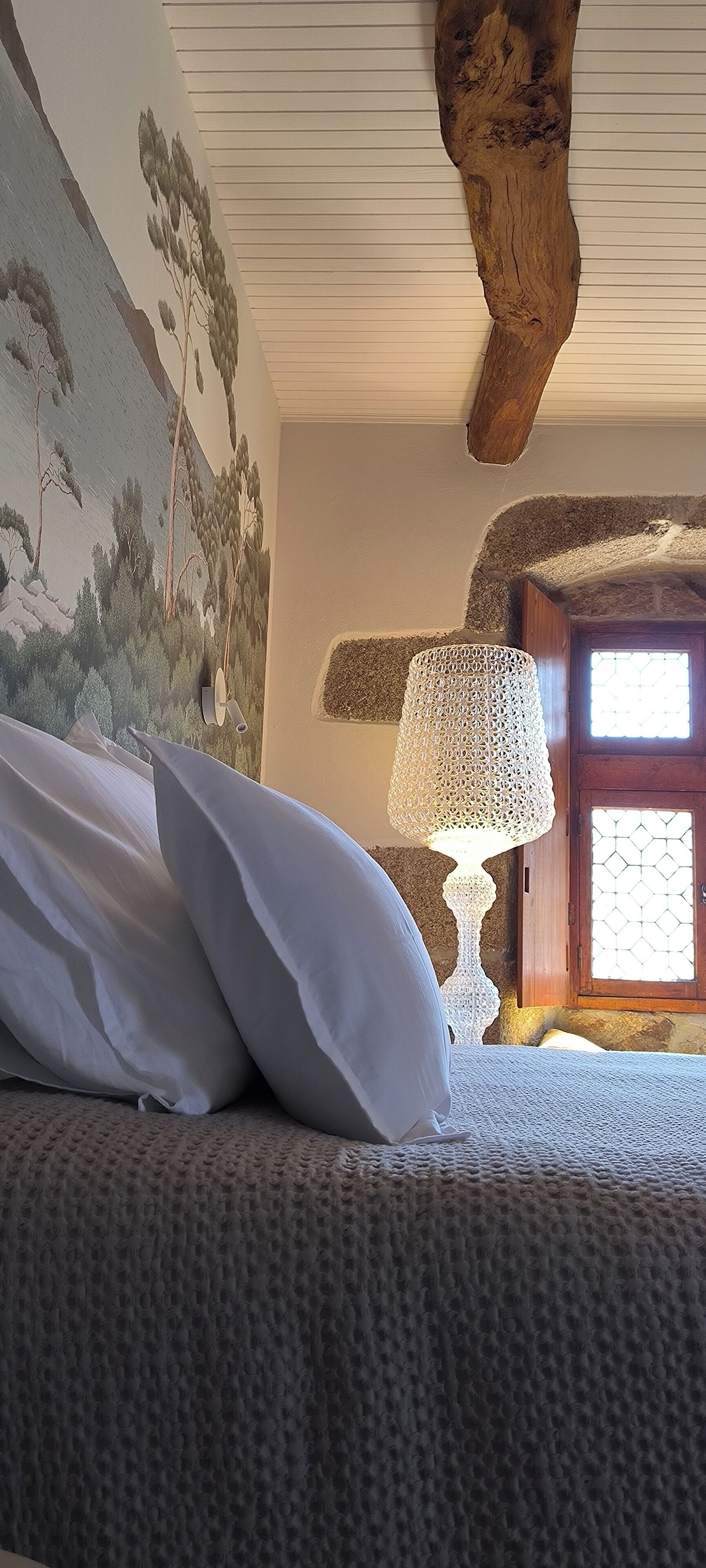 A bedroom with a bed, pillows, a textured blanket, a wall mural, a wooden ceiling beam, and a window with a wooden door.
