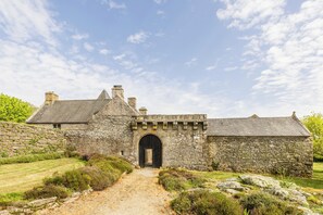 Un edificio de piedra con una entrada arqueada grande, rodeado por un muro de piedra y un sendero cubierto de césped que conduce a él.