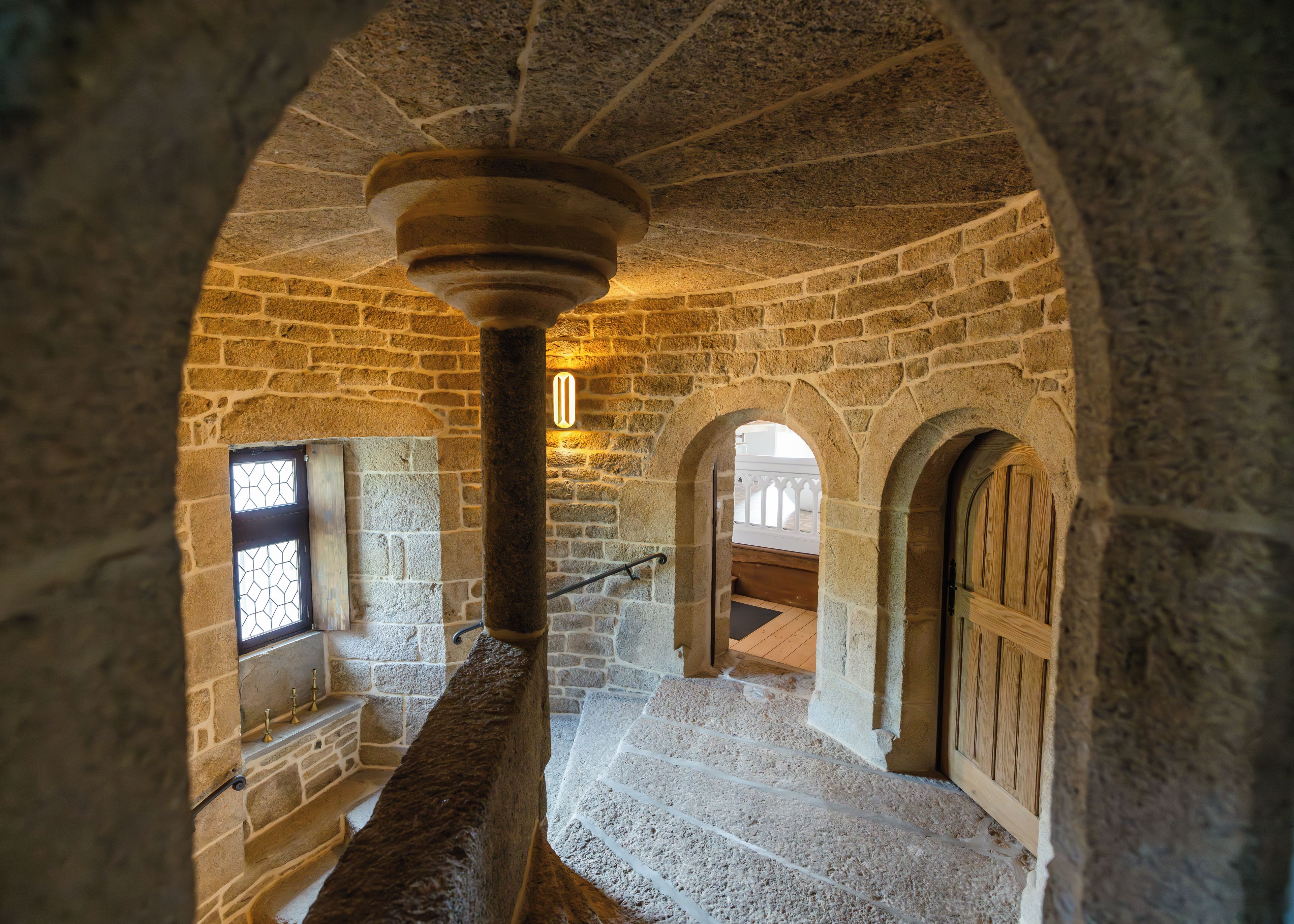 Un interior de piedra con una columna central, arcos y una escalera.