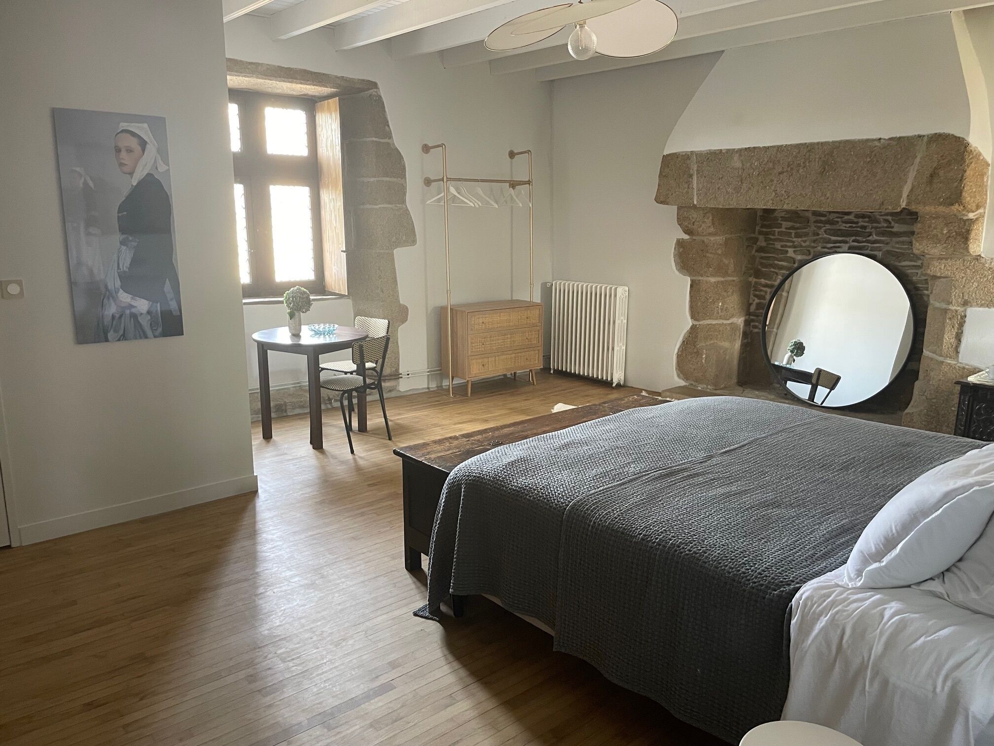 A bedroom with a bed, a round mirror, a stone fireplace, a radiator, and a small table with a chair.