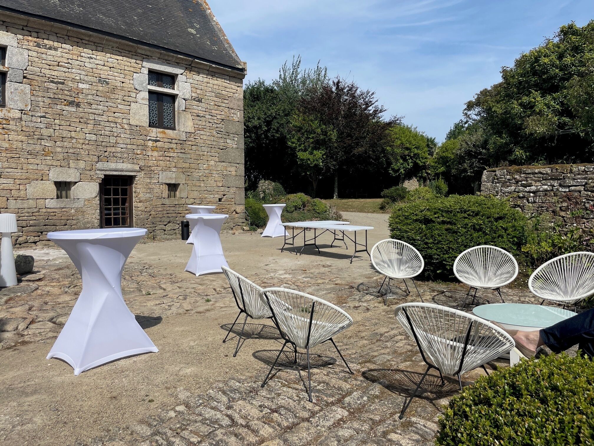 An outdoor patio area with stone tables and white chairs, a stone building in the background, and a pathway leading to it.