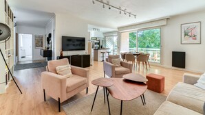 A spacious living area with a sofa, coffee table, and chairs, a kitchen in the background, and large windows offering a view of the outdoors. - Jardin des plantes - First concierge service (Montpellier)
