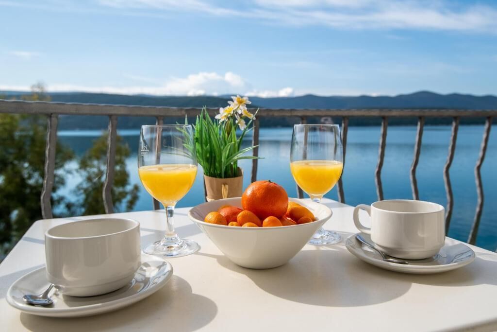Una mesa con un bol de naranjas, dos vasos de jugo de naranja y dos tazas blancas en un balcón con vista a un lago.