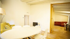Een kamer met een ronde tafel en stoelen, een bank, een televisie en een deur naar een andere kamer.