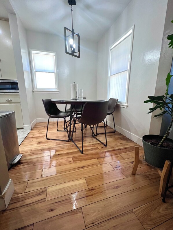 A dining area with a wooden floor, a table with four chairs, a hanging light fixture, and a potted plant.