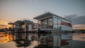 A modern floating structure with a glass facade, a lifebuoy, and a clear view of the sunset over water. - Havel River Houseboat (Zehdenick)