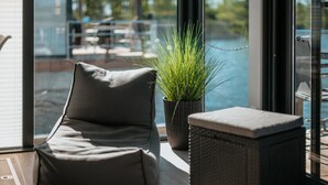 A modern room with a grey sofa, a small potted plant, and a wicker ottoman near a large window overlooking a pool. - Havel River Houseboat (Zehdenick)