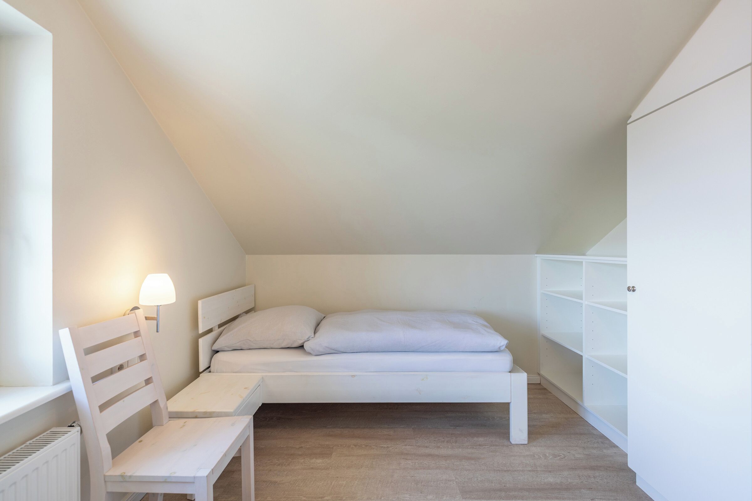 A small, white, loft-style bedroom with a single bed, a nightstand, a chair, and a lamp.