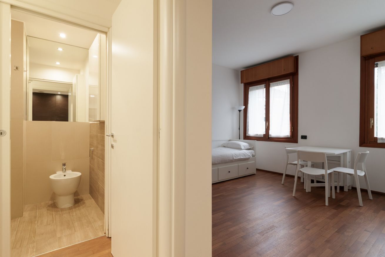 A bathroom with a toilet, a bidet, and a mirror. The room has wooden flooring and a window with curtains.