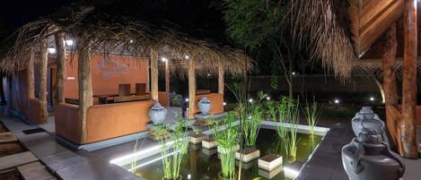 A night-time outdoor area with a thatched-roof structure, a small pool, and decorative vases.